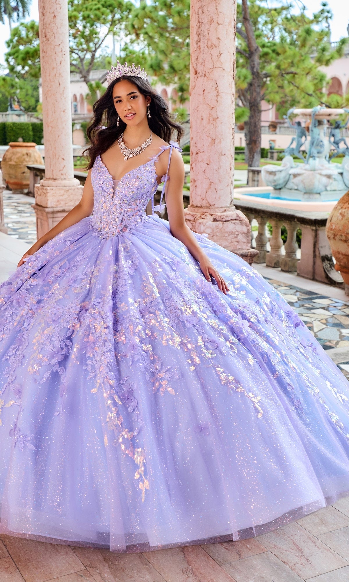 A young woman in the Quinceanera Dress PR30157 By Princesa, featuring sparkling lavender glitter and lace appliques with a tiara, stands outdoors by columns, lush gardens, and a fountain.